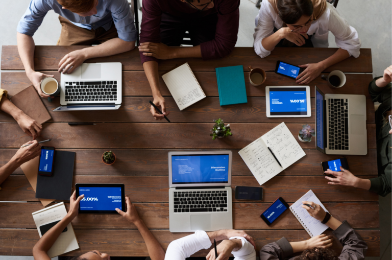 A group of co-op students sit around a table with laptops, tablets, notebooks, and smartphones, engaged collaborative work.
