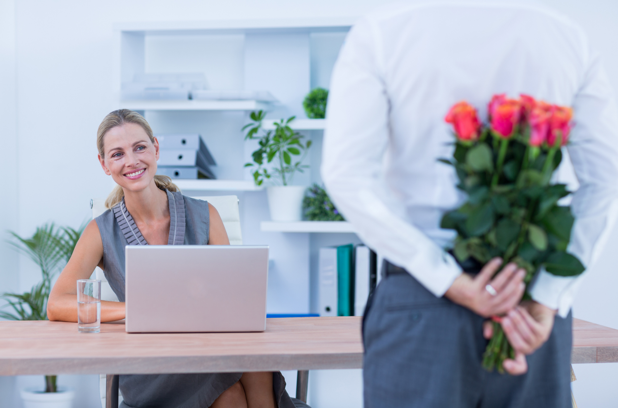 Person seated at a desk with a laptop looking at another person standing nearby holding flowers behind their back, representing workplace relationships.